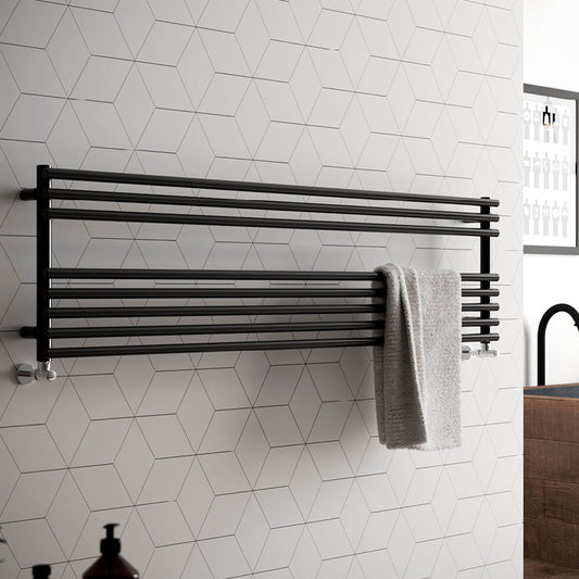 Horizontal hot water towel warmer mounted on a white tiled wall in a bathroom, with a dark grey towel hanging on it.